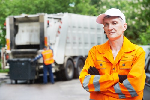 A waste collection worker stands in the foreground with arms crossed, wearing a bright orange high-visibility jacket with reflective grey and yellow stripes, a white cap, and black gloves. Behind him, a large white rubbish disposal truck with an open rear compartment is positioned on an asphalt driveway or street, with another worker operating the truck's loading mechanism. The background features lush green trees and foliage, suggesting a suburban or residential area near Euston or the broader London postcode area. The scene depicts a typical rubbish removal or clearance operation, with the truck partly loaded with waste. The ground is slightly damp, indicating recent weather activity, and natural daylight illuminates the scene evenly, emphasizing the practical setting of professional waste management work by Office Clearance Euston.