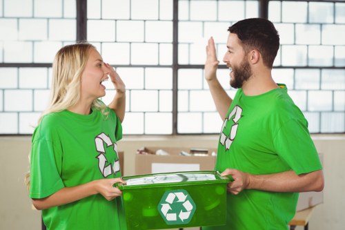 A man and a woman wearing bright green T-shirts with recycling symbols are engaged in a positive interaction indoors, in front of large industrial-style windows with a grid pattern of black metal frames and clear glass panes. The woman, with long blonde hair, is smiling broadly while touching her face, possibly in a gesture of agreement or greeting. The man, with short dark hair and a beard, is holding a transparent green recycling bin with a white recycling symbol on the front, positioned between them. The background includes stacks of cardboard boxes and a light, neutral-colored wall, suggesting an environment related to waste management or rubbish collection. The scene conveys a professional and environmentally conscious atmosphere aligned with rubbish removal services in the Euston area, with natural lighting illuminating the space evenly and emphasizing the clarity of the objects and expressions in the image.