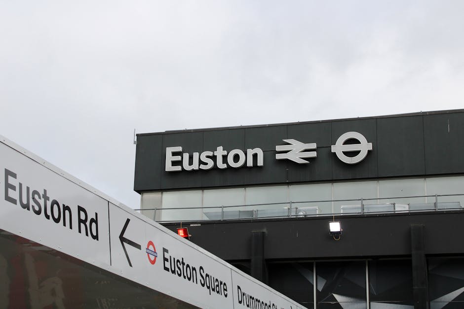 Photograph of the exterior of Euston station, displaying the station's name and the London Underground and National Rail symbols mounted on a dark grey building facade. In the foreground, a white dire