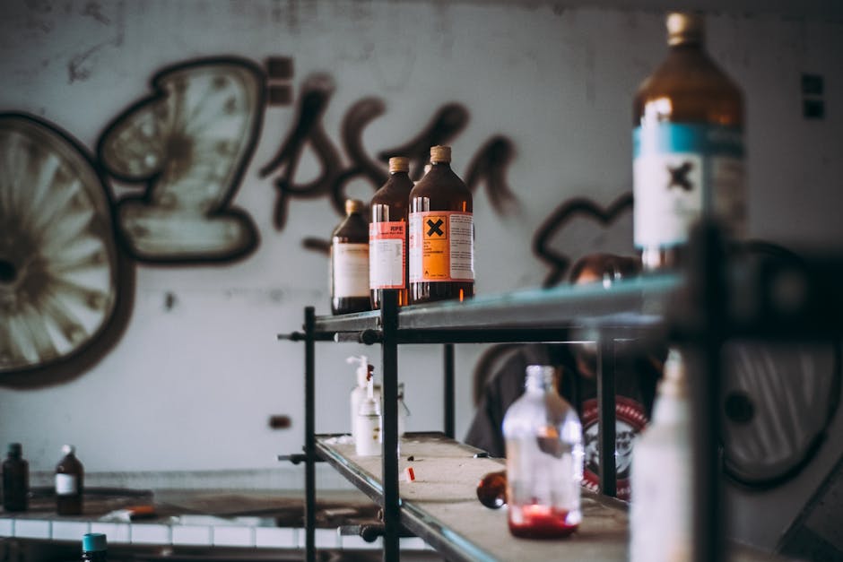 Inside a space with a metal shelving unit holding several large amber glass bottles, some with warning labels including a prominent black X symbol on an orange background. The bottles vary in size and